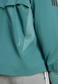 Veste teal avec une texture lisse, présentant un panneau latéral distinct et un logo sur la manche. Hém ajustable avec un cordon de serrage.