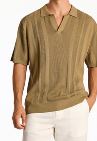 kortärmad olivgrön pikétröja med V-ringning, som har vertikal ribbad textur och subtila horisontella ränder, tillverkad av stickad tyg.