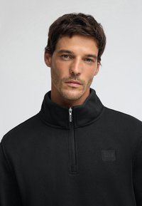 Schwarzer Zip-Up-Pullover aus strukturiertem Stoff, mit hohem Kragen und einem Reißverschluss vorne, verziert mit einem dezenten quadratischen Logopatch.