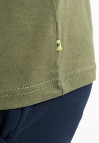 Oliwkowy bawełniany t-shirt z krótkimi rękawami i bocznym szwem, z małą żółtą metką, w połączeniu z granatowym materiałem poniżej.