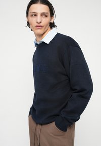 Pulover tricotat bleumarin cu guler rotund, având un logo embossat în față, purtat peste o cămașă cu guler albăstruie, asortat cu pantaloni maro.