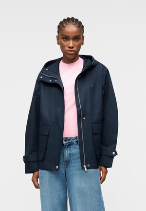 Kobieta z zaplecionymi włosami ubrana w granatową kurtkę z kapturem na różowym swetrze i jasnoniebieskich szerokich dżinsach, stojąca na jednolitym tle.