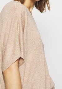 Beżowy sweter dzianinowy z krótkimi rękawami, luźny krój i subtelna struktura. Materiał wydaje się miękki i lekko melanżowy.