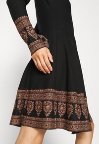 Robe noire à manches longues ornée d'une broderie florale complexe de couleur cuivre sur les poignets et l'ourlet, portée par une personne debout.