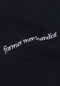 Svart stoff med hvit brodert tekst som sier "former merchandise." Teksturen ser glatt og myk ut, uten mønstre eller aksenter.