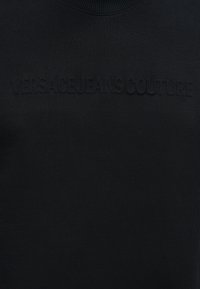 Μαύρο ύφασμα με ανάγλυφο μαύρο κεντημένο κείμενο που αναγράφει "VERSACE JEANS COUTURE" στην περιοχή του στήθους.