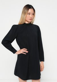 Abito nero con collo alto, maniche lunghe a sbuffo e vestibilità rilassata. Realizzato in un tessuto leggero con una texture morbida e una leggera lucentezza.