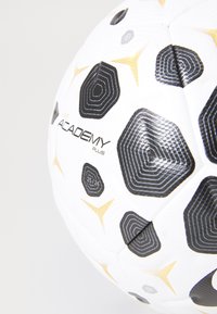 Hvid fodbold med sorte og gyldne geometriske mønstre og "Nike Academy Plus" brand på en lys baggrund.