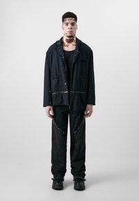 Schwarze Stoffjacke mit fransigen Kanten, ausgestattet mit Taschen und silbernem Zubehör. Kombiniert mit abgenutzten schwarzen Hosen und klobigen schwarzen Schuhen.