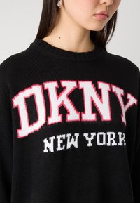 Černý pletený svetr s velkým bílým a růžovým nápisem "DKNY" a "NEW YORK" v bílé barvě, s ribbovaným výstřihem a hladkou texturou.