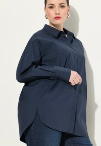 Camisa de botón azul marino con cuello, mangas largas y dobladillo curvado. Tejido suave con un ligero brillo y pequeñas aberturas laterales.