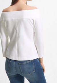 Schulterfreies weißes Oberteil mit Dreiviertelärmeln, glatter Textur, figurbetontem Design, kombiniert mit blauen Jeans aus Denim mit schrägen Gesäßtaschen.