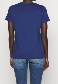 Osoba oblečená v jednoduchém krátkém rukávu navy modrého trička a středně modrých džínách, zobrazená zezadu na světlém pozadí.