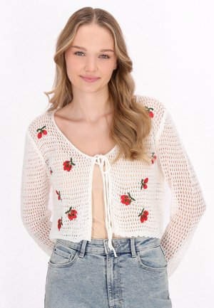 Weißer gestrickter Cardigan mit einem cropped Design, verziert mit roter floraler Stickerei und einer Bindeschließe. Über einem beigen Oberteil getragen, kombiniert mit heller Denim.