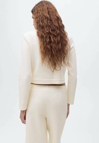 Blazer crema tagliato con maniche lunghe, texture liscia e una vita marrone, abbinato a pantaloni a gamba larga coordinati.