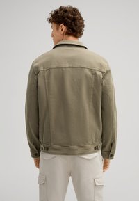 Giacca in denim khaki con colletto classico, polsini con bottoni e vestibilità rilassata. Dettagli di cucitura notevoli e pantaloni cargo bianchi.