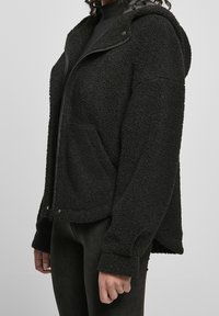 Personne portant une veste à capuche zippée noire texturée avec des poches, associée à un pantalon noir sur un fond gris uni.