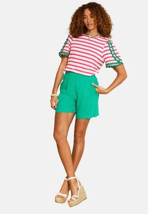 Žena s kudrnatými vlasy, oblečená v růžovo-bílém pruhovaném tričku se zeleným lemováním, zelených šortkách a bílých klínových sandálech, stojící s rukou v bok.