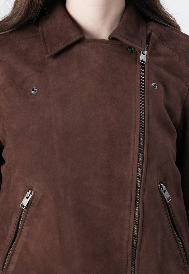 Brązowa zamszowa kurtka z zapięciem na suwak z przodu, z kołnierzem zapinanym na napy i srebrnymi detalami. Posiada boczne kieszenie na suwak oraz gładką teksturę.