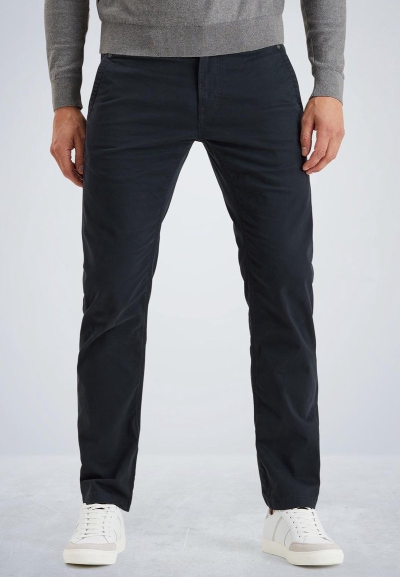 Homme portant un pantalon noir slim, un pull gris et des baskets blanches, debout sur un sol gris clair devant un fond uni.