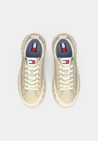 Sneakers beige con suole in gomma testurizzata, lacci bianchi e una soletta blu con un'etichetta logo rosso, bianco e blu sul lato.