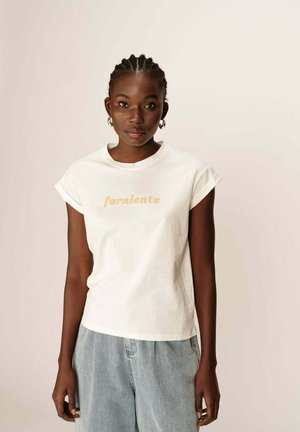 T-shirt en coton blanc avec des manches courtes retroussées, portant "farniente" imprimé en marron clair. Porté avec un short en denim bleu clair.