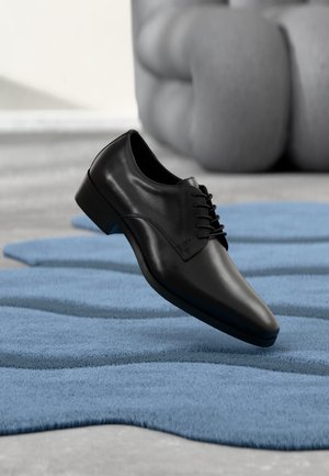 Chaussure habillée en cuir noir poli suspendue au-dessus d’un tapis bleu texturé avec un canapé gris flou en arrière-plan.