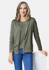 Olijfgroen vest met een ronde hals, ribbelstructuur, sluiting met knopen aan de voorkant en lange mouwen. Gedragen over een bijpassend topje en blauwe spijkerbroek.