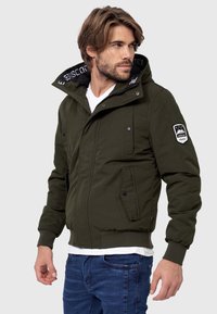 Giacca bomber verde oliva con zip frontale, chiusure a scatto e cappuccio. Presenta due tasche anteriori e una patch sulla manica sinistra.