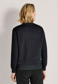 Vrouw met kort blond haar, gekleed in een gestructureerde zwarte trui en zwarte broek, die met de rug naar de camera staat tegen een effen lichte achtergrond.