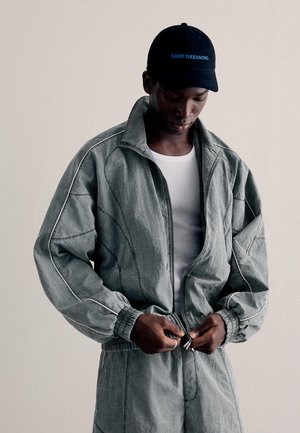 Férfi szürke cipzáros dzsekiben és hozzáillő nadrágban, fehér ingben, fekete sapkával, amin a "KEEP DREAMING" felirat olvasható, lefelé néz, miközben begombolja a dzsekit.