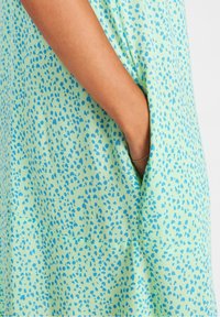 Jasnozielona sukienka z fakturowanym wzorem małych niebieskich kropek, z widoczną boczną kieszenią i miękkim składem materiału.