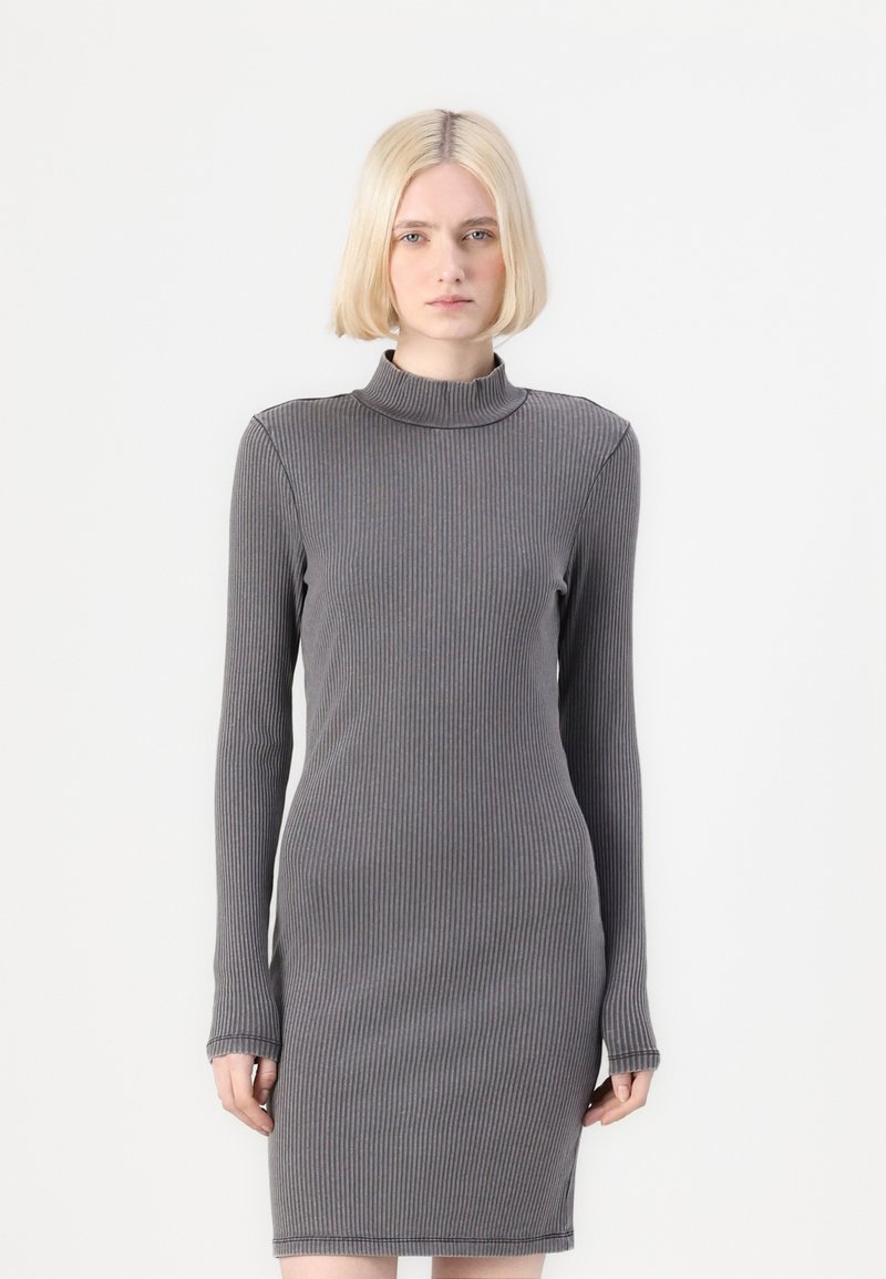 Ξανθιά γυναίκα φοράει ένα στεντό, ριμπ τούλινο φόρεμα σε σκούρο γκρι χρώμα με ψηλή λαιμόκοψη, στέκεται μπροστά σε απλό λευκό φόντο.