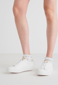 White athletic shoes with a smooth texture, featuring a minimalist design and light brown accents. Worn with frilled white ankle socks.