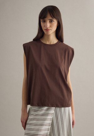 Jonge vrouw met steil bruin haar draagt een mouwloze bruine top met gewatteerde schouders en een gestreepte witte en bruine rok tegen een effen achtergrond.