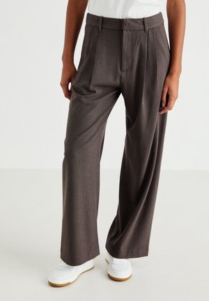 Pantalon marron à jambes larges à carreaux porté avec des baskets blanches et un haut blanc, montré du milieu du torse aux pieds sur un fond neutre.