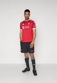 Punane Liverpool FC särk valgete detailide ja mustade lühikeste pükstega ning mustade jalgpallijalatsitega. Lühikeste varrukatega disainil on logod ja sponsorid.