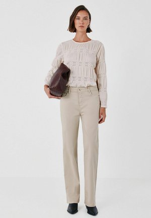 Femme portant un chemisier en dentelle crème et un pantalon beige, tenant une grande pochette texturée marron foncé, debout devant un fond blanc uni.