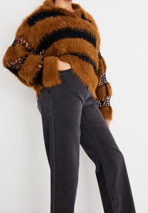 Persoon draagt een bruin-zwart gestreepte, pluizige trui met opengewerkte mouwen en donkergrijze jeans met hoge taille, handen in de zakken.