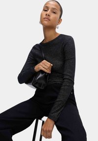 Schwarzes Langarmoberteil mit schimmernder Textur, kombiniert mit einer kleinen schwarzen Tasche. Das Modell trägt dunkle Hosen und große silberne Ohrringe.