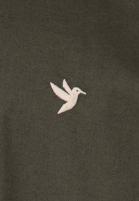 Olivgrüner Stoff mit subtiler Textur, versehen mit einem weißen, gestickten Kolibri-Logo, das auf der linken Seite platziert ist.