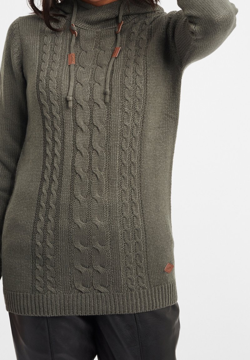 Personne portant un pull en maille torsadée vert olive avec une capuche et des cordons, associé à un pantalon en cuir noir.