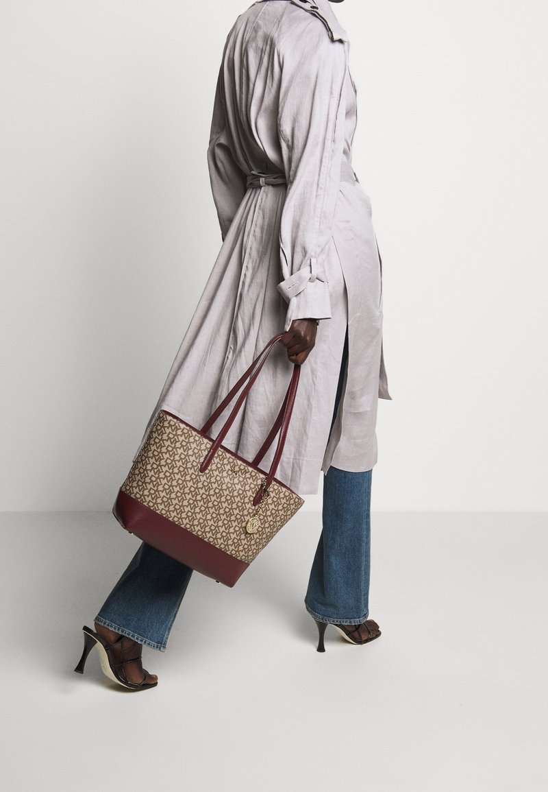 Personne en jean bleu et trench beige marchant, portant un sac fourre-tout beige et bordeaux à motifs, avec des doubles poignées.