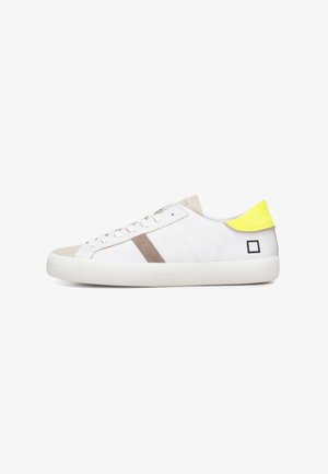 Zapatilla blanca de caña baja con acentos beige y gris, lengüeta del talón en amarillo brillante, suela blanca y pequeño logo cuadrado negro cerca del talón sobre un fondo liso.