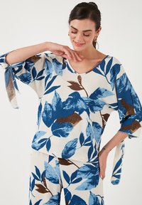 Top blu e bianco con motivo fogliare, scollatura a V, maniche a tre quarti e lacci sulle maniche. Il tessuto ha una finitura testurizzata.