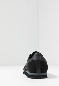 Svart Asics-sneaker sedd bakifrån, som visar nät- och läderpaneler med en grå och svart sula på en vit yta.