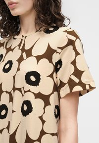 Nærbilde av en person som har på seg en beige og brun blomster-mønstret kortermet topp med store svarte midtpunkt blomster.