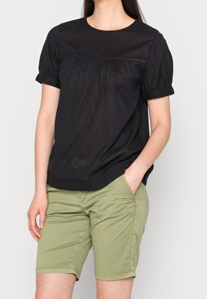 Kvinde iført en sort kortærmet bluse og lysegrønne knælange shorts med den ene hånd i lommen mod en ensfarvet baggrund.