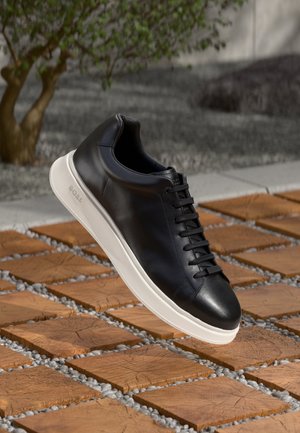 Czarny skórzany sneaker z białą podeszwą oznaczony napisem „BOSS”, unoszący się nad drewnianymi i żwirowymi płytkami na zewnątrz, w pobliżu rozmytego drzewa.