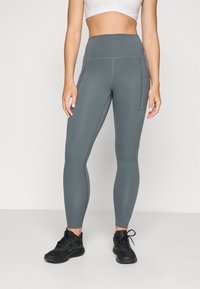 Leggings de yoga taille haute, gris avec des coutures flatlock et des poches latérales, associés à des chaussures de sport noires. Tissu extensible, design élégant.